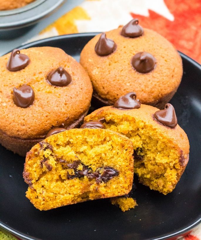 three pumpkin chocolate chip muffins on a black plate, with one muffin cut in half to show the chocolate chips inside