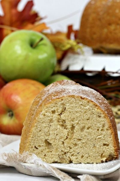 Slice of apple cider bundt cake on a white plate with apples in the background