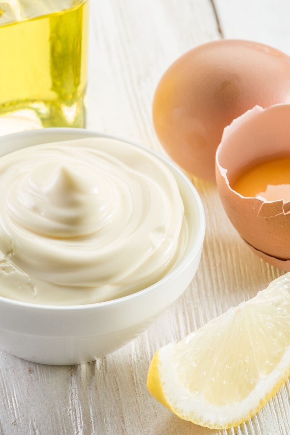 a white bowl of mayonnaise next to eggs and oil and a lemon wedge