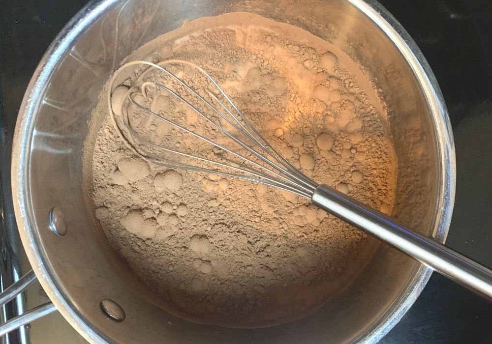 cocoa powder, oat milk, and sugar combined in a saucepan, with a whisk in the pan. The cocoa powder has not yet dissolved into the milk.