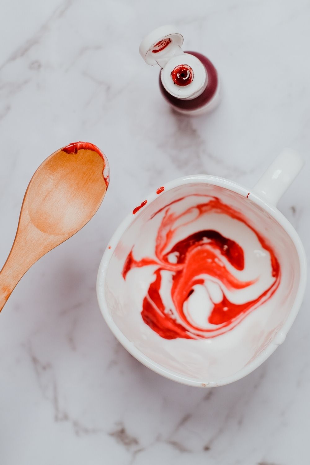 a bottle of red gel food coloring next to a bowl of white icing, where the food coloring is being swirled into the icing.