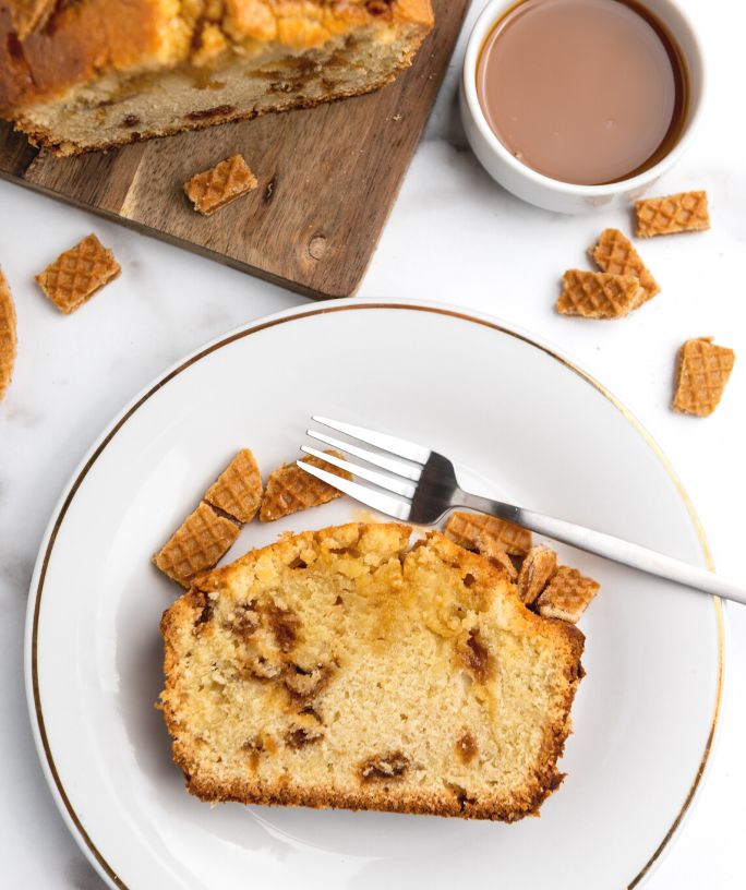 overhead view of a slice of stroopwafel cake served on a white plate, with coffee and the remaining cake in the background