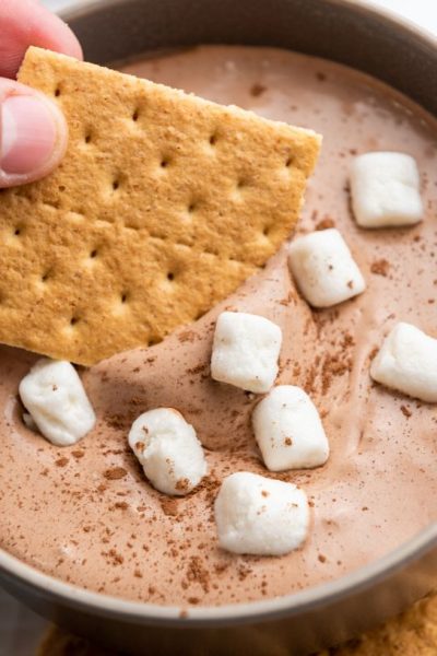 a woman's hand dips graham crackers into hot cocoa dip