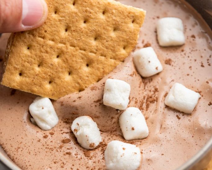 a woman's hand dips graham crackers into hot cocoa dip