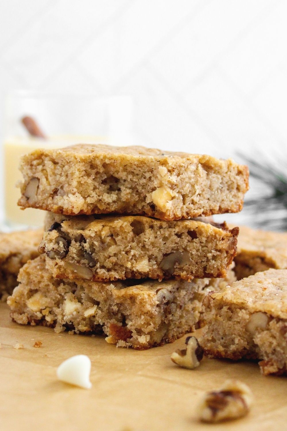 a few eggnog blondies stacked together, with walnuts and chocolate chips scattered around and a glass of eggnog in the background