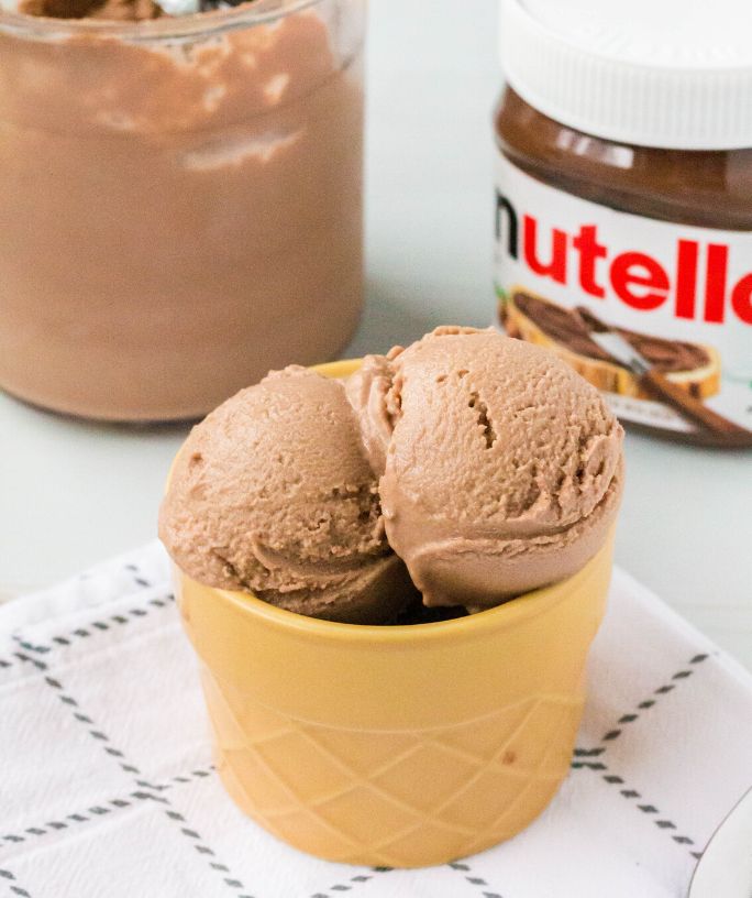a dessert cup holds a couple of scoops of Ninja Creami Nutella ice cream. A jar of Nutella is in the background, along with a Ninja Creami pint.