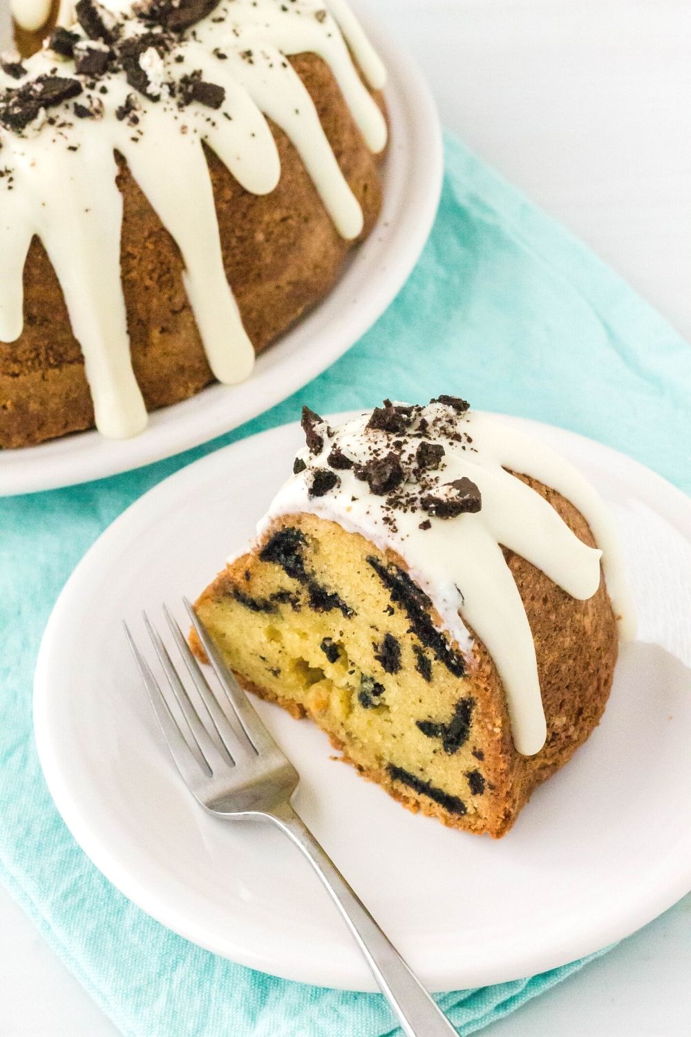 a slice of pound cake with oreos served on a white plate, with a fork alongside it and the rest of the cake in the background