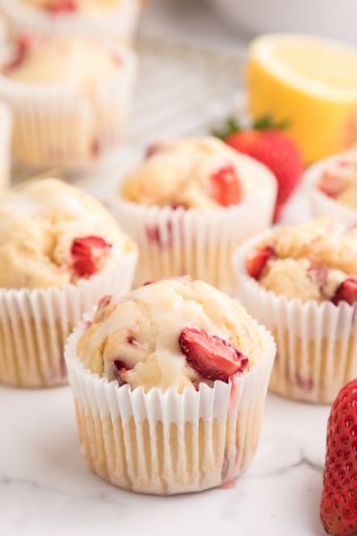a few homemade strawberry lemon muffins on a marble counter, with a fresh strawberry and half of a lemon in the view as well