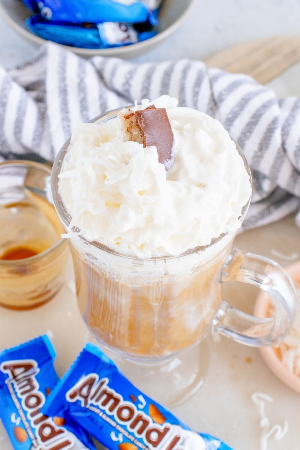 overhead view of a glass mug of almond joy coffee, topped with whipped cream and coconut shreds, with almond joy candy bars scattered around