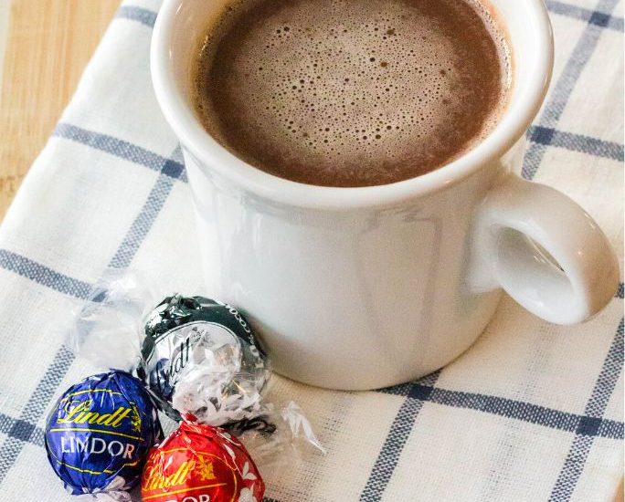 a white mug of Lindt hot chocolate rests on a blue and white napkin, with three Lindor truffles next to it.