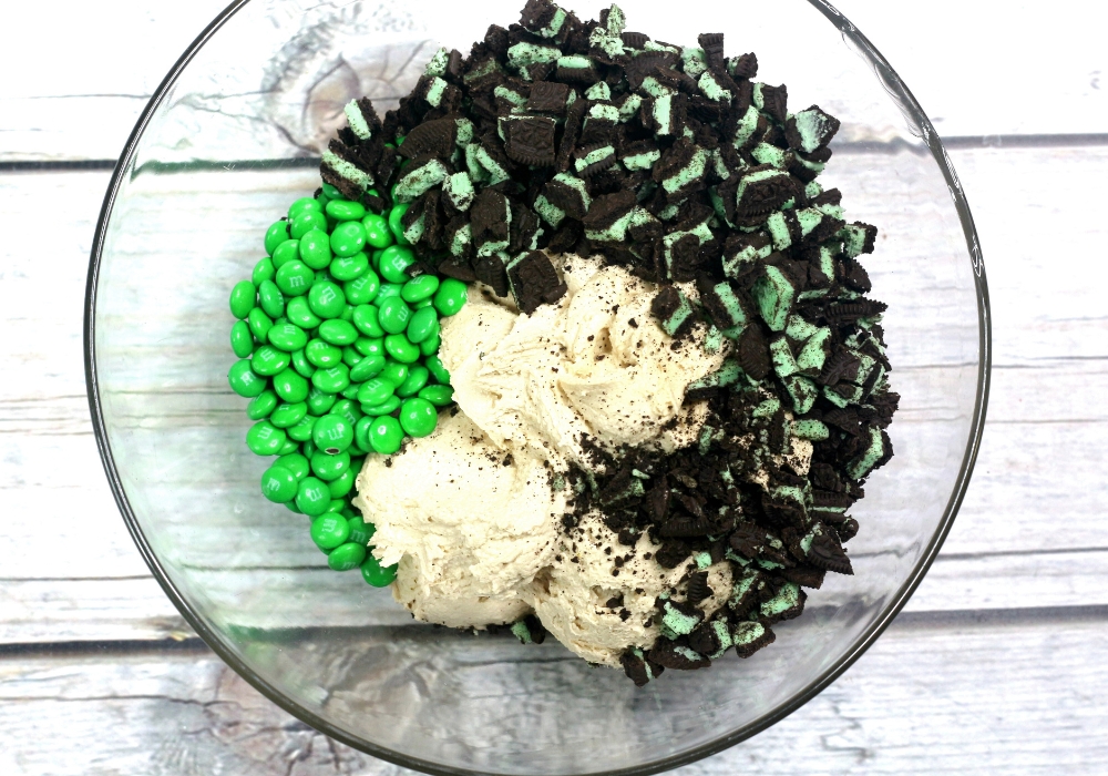 overhead view of a mixing bowl filled with cookie dough, green M&Ms, and mint Oreo cookie pieces.
